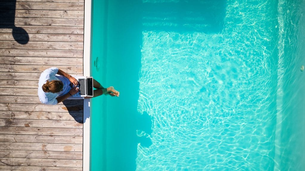 A top-down view of a person working on a laptop beside a bright blue pool, illustrating modern pool marketing strategies for 2025