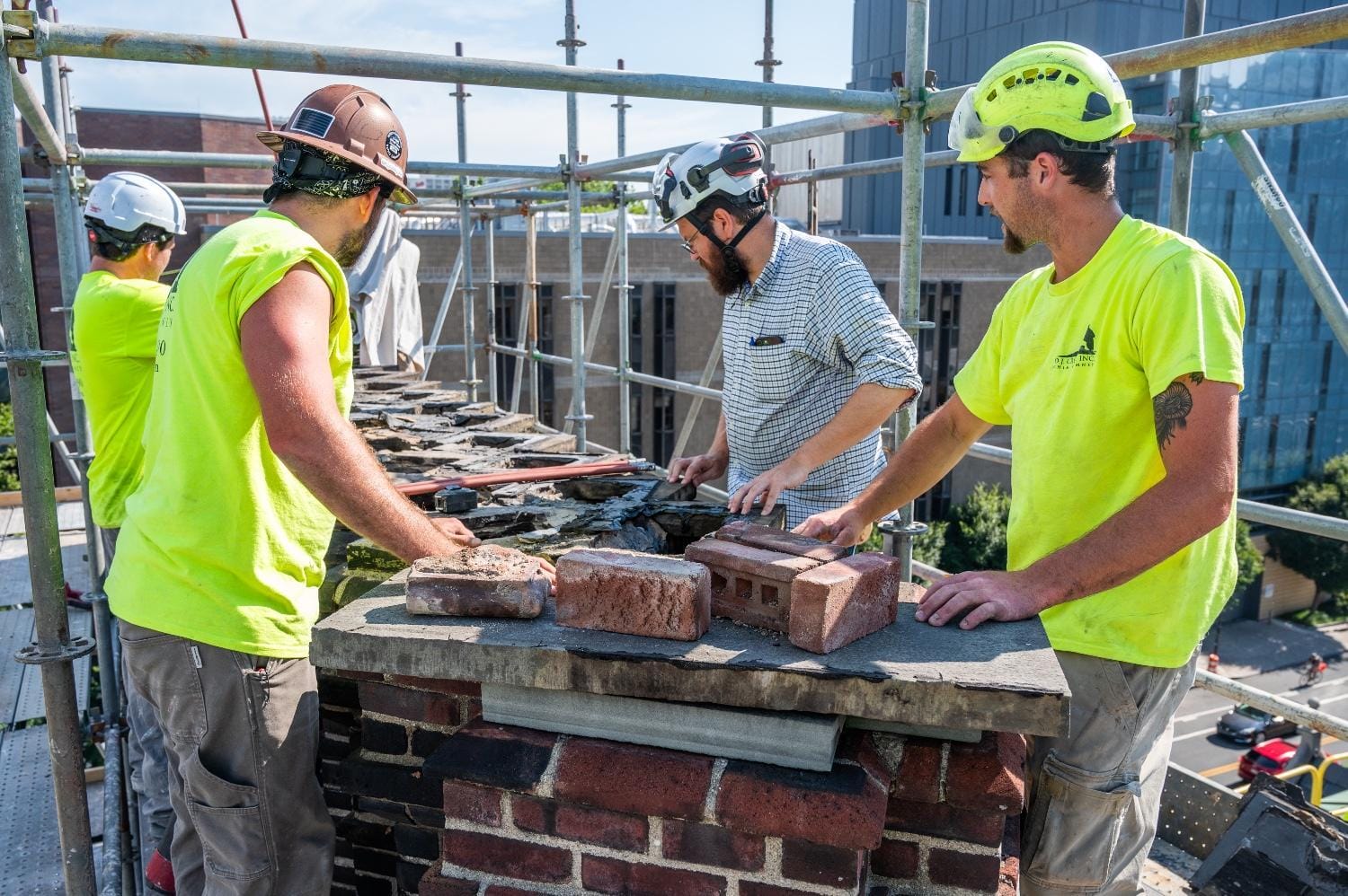 Original infographic shows four construction workers wearing safety helmets and high-visibility shirts standing on scaffolding while repairing the top of a brick chimney. Two workers in bright yellow shirts are on either side of the chimney, while another in a brown helmet and one in a plaid shirt are at the center applying mortar and positioning loose bricks. Several red bricks and trowels rest on the chimney ledge, and a cityscape with buildings and streets can be seen in the background under a clear blue sky.