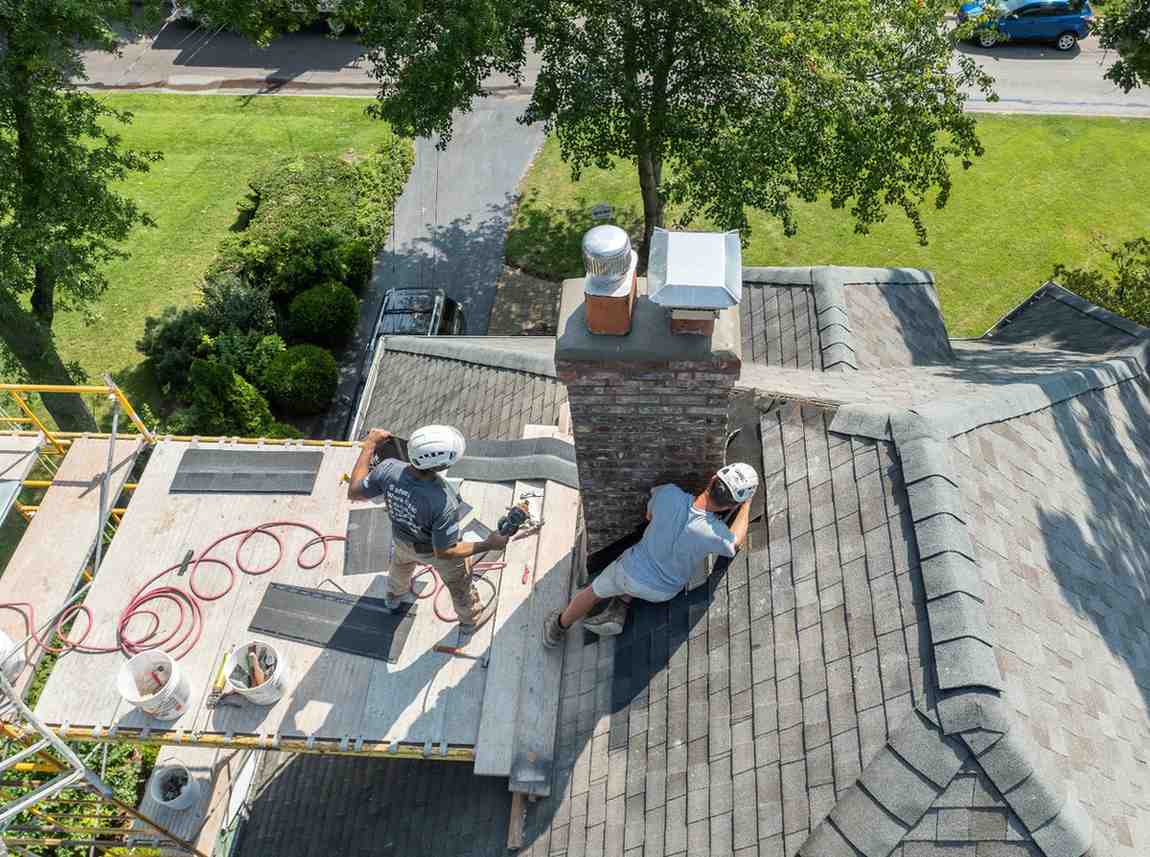 Original infographic shows two workers wearing helmets and gray shirts repairing or inspecting a brick chimney on top of a shingled residential roof. They are surrounded by scaffolding and safety equipment, with tools, hoses, and buckets visible on the wooden platform. One worker is standing on the scaffolding using a tool, while the other is kneeling on the roof near the chimney base. The scene is brightly lit with green trees and a suburban street visible below, emphasizing a clear, sunny day.